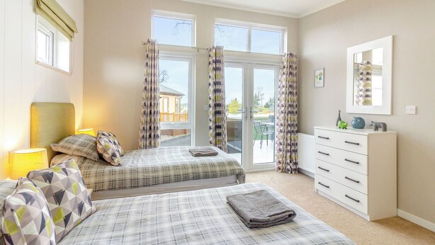 A bedroom with two beds, a dresser, a mirror, and a view through a glass door. - Luxury modern 2 Bedroom Lodge. (Swinethorpe, Nr Lincoln)