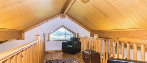 Interior de madera con techo inclinado, una ventana, un sofá de cuero negro, un cofre de madera y un tapete estampado.