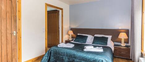 Un dormitorio con puerta de madera, cama con ropa de cama color turquesa, dos lámparas de noche y una mesita de noche.