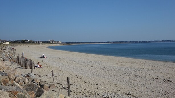 Une vaste plage de sable fin, un ciel bleu dégagé, une clôture en bois et quelques personnes assises sur le sable.