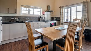 A modern kitchen with a wooden dining table and chairs, a built-in oven, and a window with curtains. - 4 bedroom accommodation in Hornsea (Hornsea)