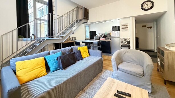 Una sala de estar moderna con un sofá, una butaca, una mesa de centro de madera, una escalera y una zona de cocina.
