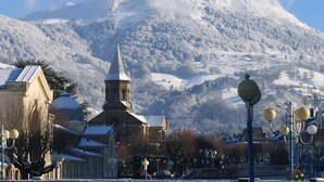 Un pueblo con una iglesia que destaca por su alta aguja, rodeado de edificios cubiertos de nieve y una montaña al fondo.