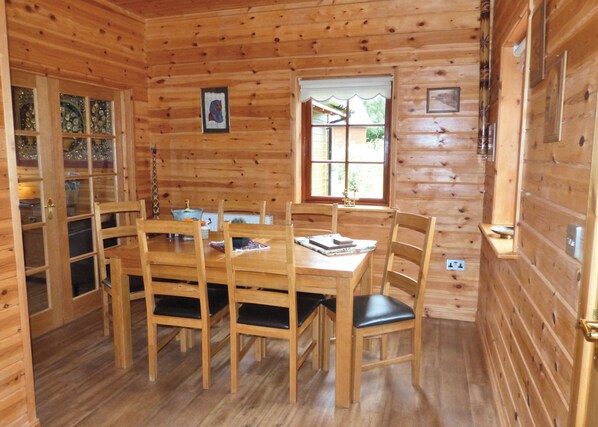 A wooden cabin interior with a dining table, chairs, a window, and a door with glass panels.