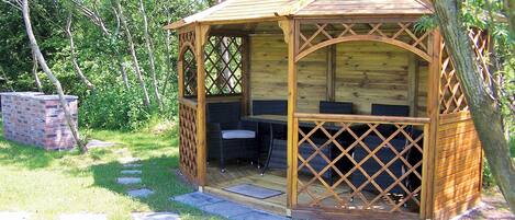 A wooden gazebo with a lattice roof, outdoor seating, and a brick structure in the background.