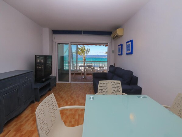 A living room with a TV, a sofa, a table, and a balcony with a view of the sea.