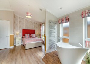 A bedroom with a bed, bedside tables, a red headboard, a window with curtains, a bathroom with a bathtub, and a wooden floor.