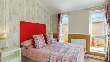 A bedroom with a red headboard, a bed with plaid bedding, a bedside table with a lamp, and a window with curtains.