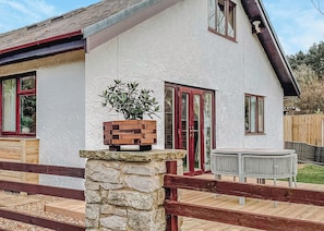 Una casa con un pilar de piedra, una valla de madera, una planta en maceta, una puerta roja y una mesa redonda.