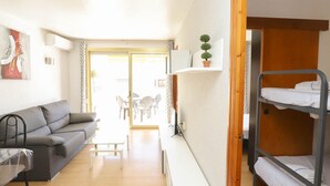 Un espacio habitable compacto con un sofá gris, una mesita, una cama litera y un ventanal corredizo que da a un área exterior con sillas y mesas.