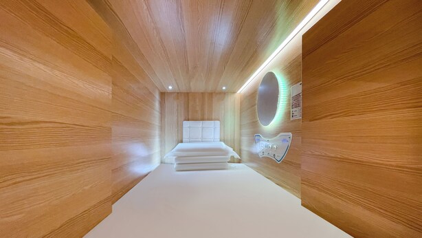 A modern hotel room with wooden panel walls, a central bed, a round mirror with LED lighting, and a control panel on the wall. - Airone Capsule Hotel (Hobart)