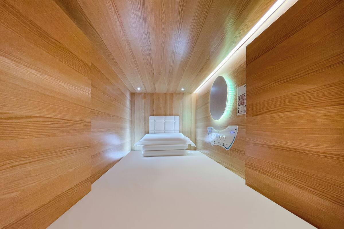 A modern hotel room with wooden panel walls, a central bed, a round mirror with LED lighting, and a control panel on the wall.