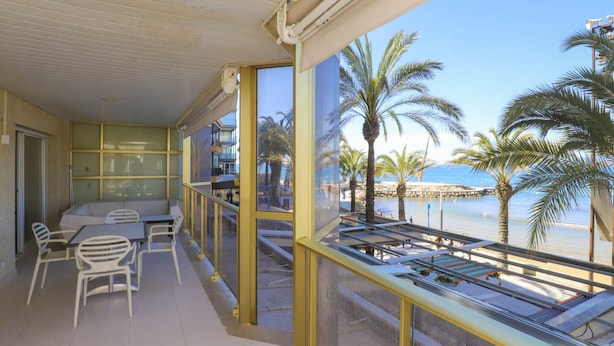 Un balcón con vistas a la playa, sillas blancas y una mesa.