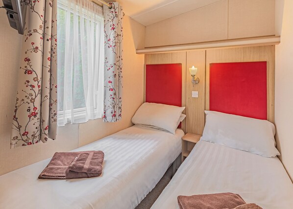 Habitación doble con dos camas, una ventana con cortinas traslúcidas, una lámpara fijada a la pared y un cabecero de madera.