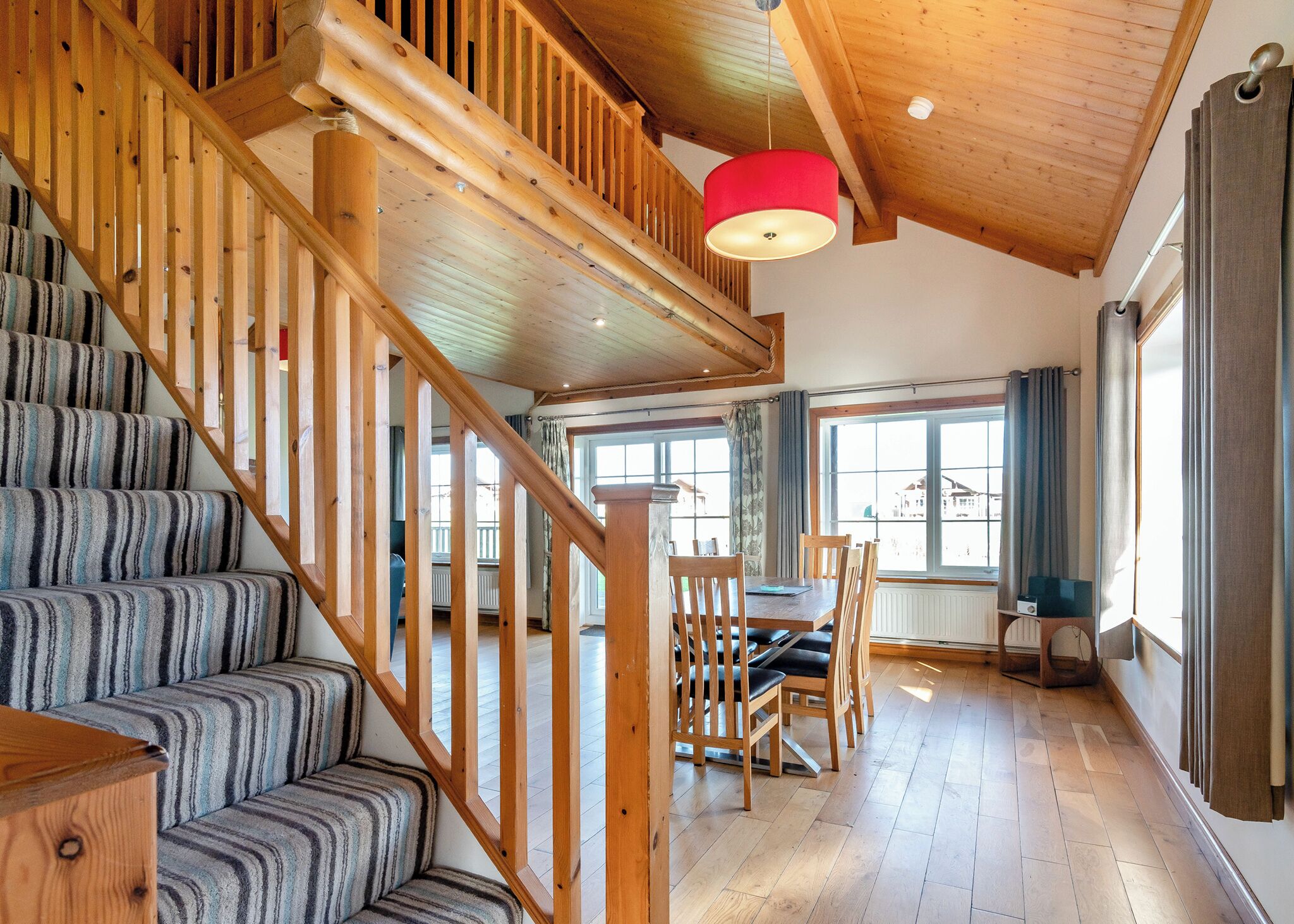 A wooden staircase with striped carpet, a dining area with a table and chairs, and large windows with curtains.