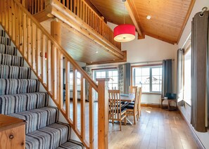 Una escalera de madera con alfombra a rayas, un comedor con mesa y sillas, y ventanas grandes con cortinas.