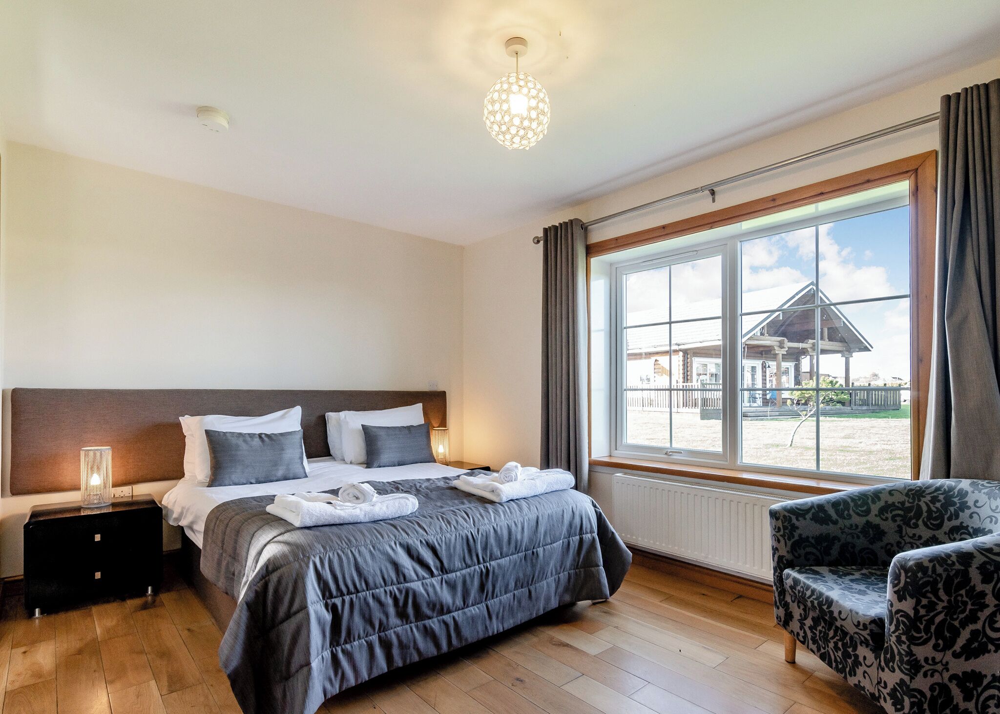 Habitación de hotel con cama, mesita de noche, lámpara, silla, ventana grande y vistas al exterior.