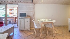 Uma sala de jantar com uma mesa e cadeiras brancas, uma televisão sobre um móvel e vista para edifícios lá fora.