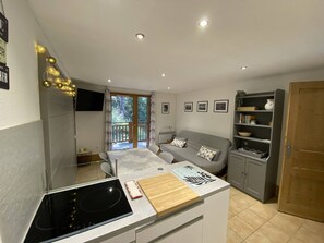 Une cuisine compacte dotée d’une cuisinière intégrée, un coin repas avec une table et des chaises, un canapé gris et une porte en bois.