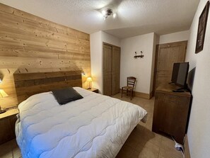 A bedroom with a wooden headboard, a bed with white bedding, a chair, a television on a cabinet, and a wall-mounted light fixture. - Cozy Apartment 6 Guests, Balcony, Pool Access, Close to Slopes and Amenities (La Plagne-Tarentaise)