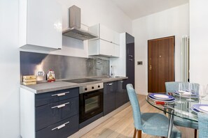 A modern kitchen with dark cabinets, a stainless steel range hood, a built-in oven, and a glass dining table with chairs.