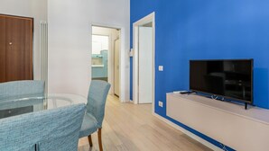 A modern living room with a glass table, blue walls, and a flat-screen TV. - GuestHost - Cozy renovated 52sqm apartment located on the first floor of a Milanese building with lift (with 4 steps to climb at the entrance). The property is located in a quiet and residential neighborhood, but at the same time a few steps from the Cen (Milano)