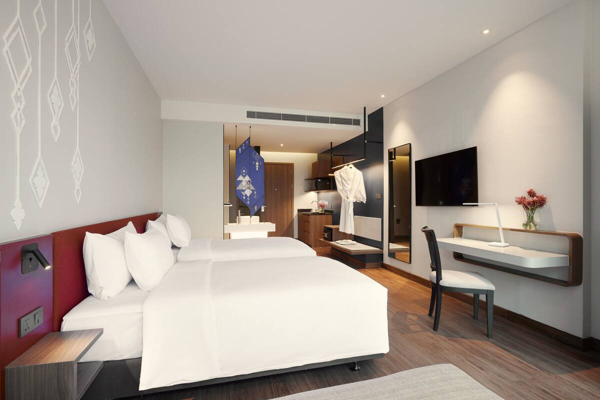 Ein modernes Hotelzimmer mit einem großen Bett, einem Schreibtisch, einem Sessel, einem Fernseher und einem im Hintergrund sichtbaren Badezimmer.