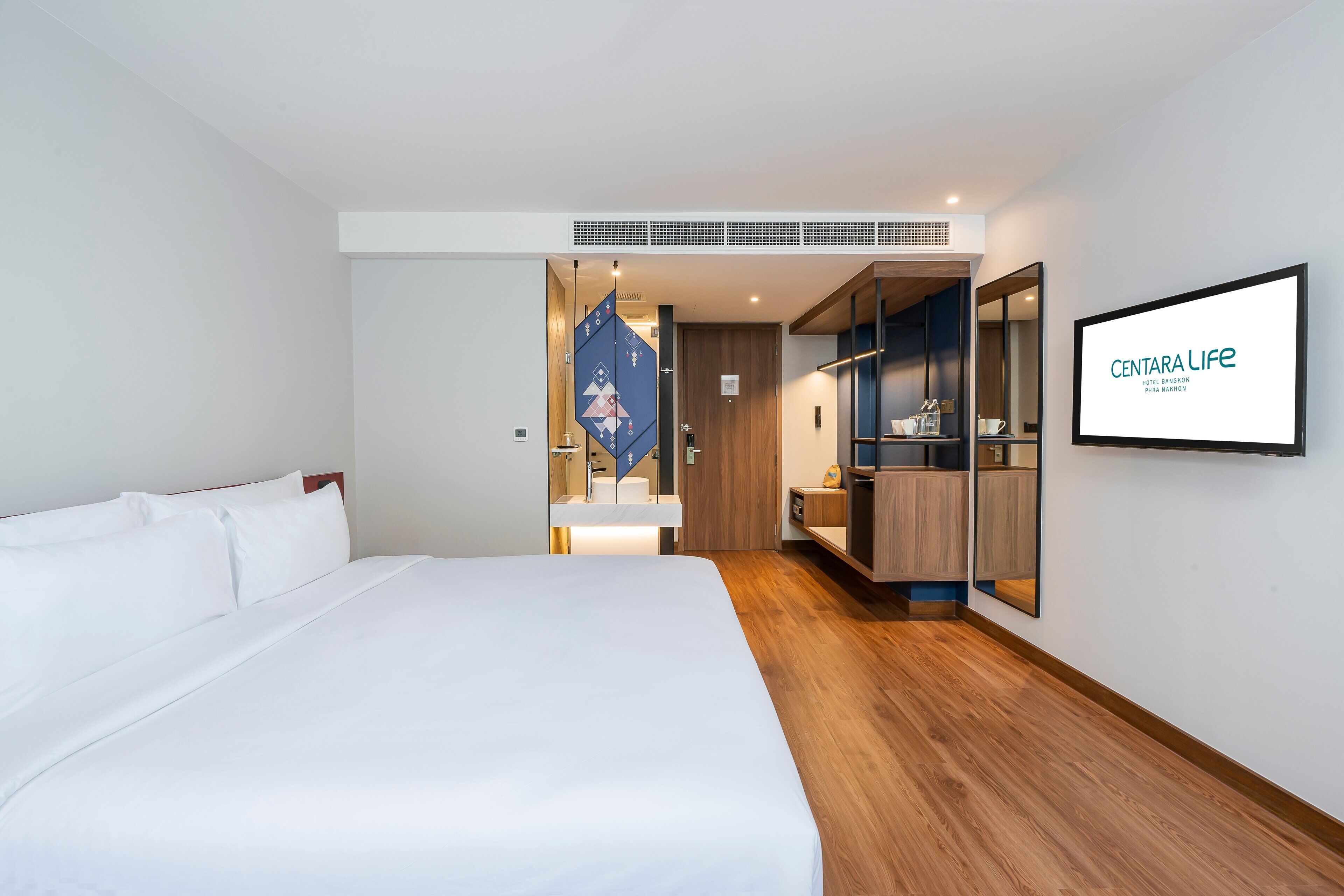 Habitación de hotel moderna con una cama grande, un armario, un espejo y un televisor que muestra el nombre del hotel.