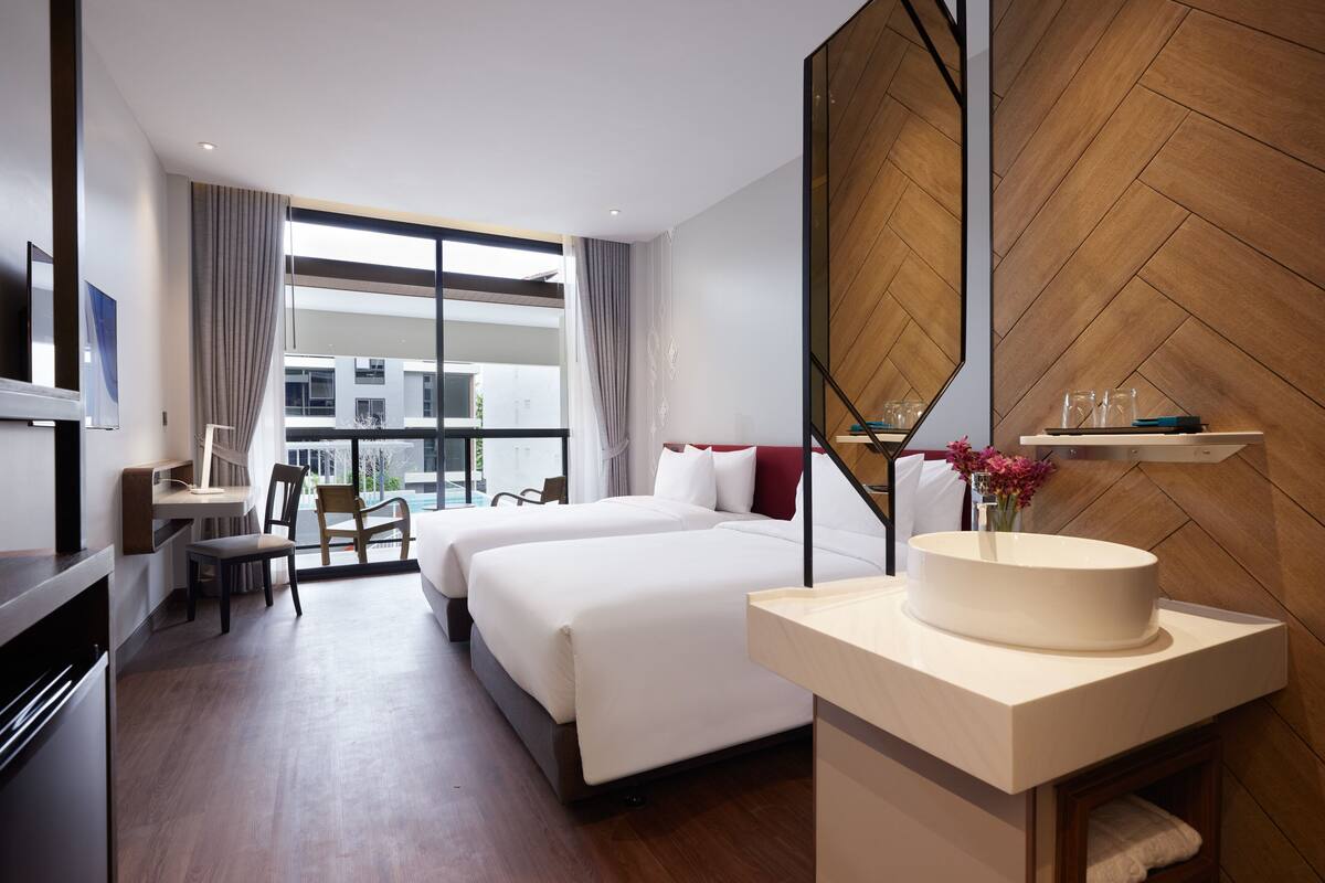 Ein Hotelzimmer mit zwei Betten, einem Schreibtisch, einem Fernseher und einem großen Fenster mit Blick auf Gebäude.