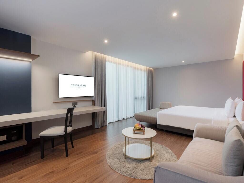 Una habitación de hotel moderna con televisión de pantalla plana, sofá, cama, escritorio con silla y una pequeña mesa redonda con frutero.