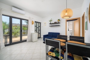 A modern living room with a blue sofa, a dining table with chairs, a small refrigerator, and a large window with a view of greenery. - Apartment Panous 2 (Stranici kod Nove Vasi)