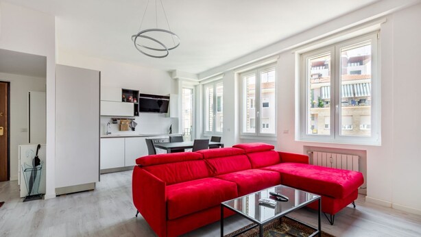 A modern living room with a red sectional sofa, a glass coffee table, and a view of the city through large windows. - Cozy and bright 66sqm apartment located on the 6th floor of a building with lift (with a flight of stairs at the entrance to reach it).The accommodation is located in an exceptional position, just 300m from P.zza Gae Aulenti, famous for the wonderful vie (Milano)