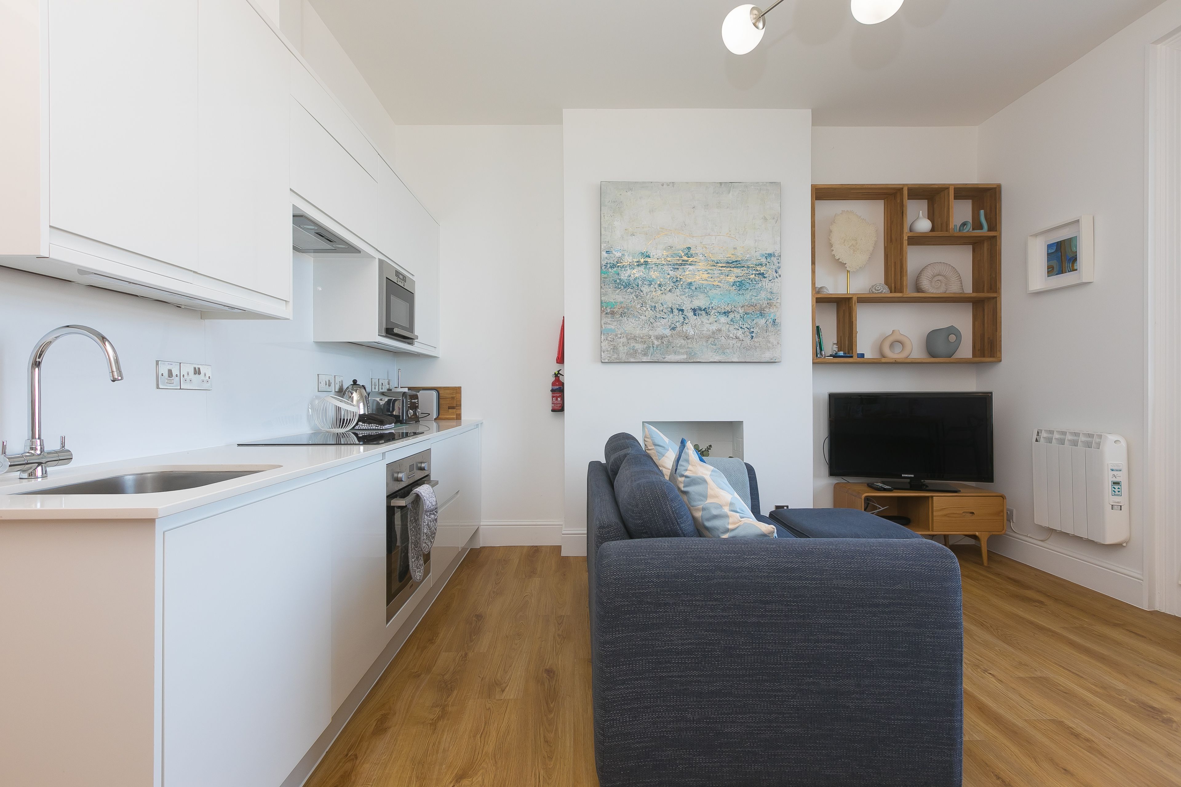 Una cocina moderna con armarios blancos, un lavamanos de acero inoxidable y un microondas. En la zona de estar, un sofá gris con cojines azules y amarillos, un televisor de pantalla plana y una estantería con objetos decorativos.