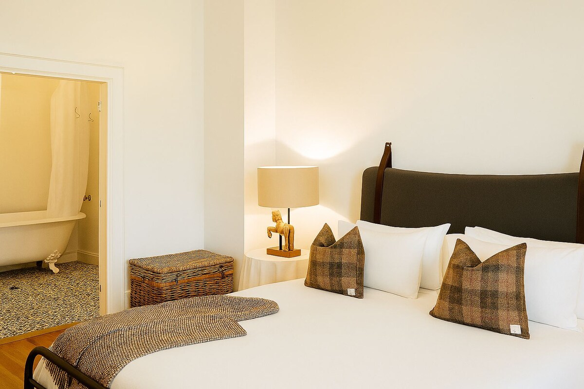 A hotel room with a bed, a bedside table with a lamp, a wicker chest, and a chair.