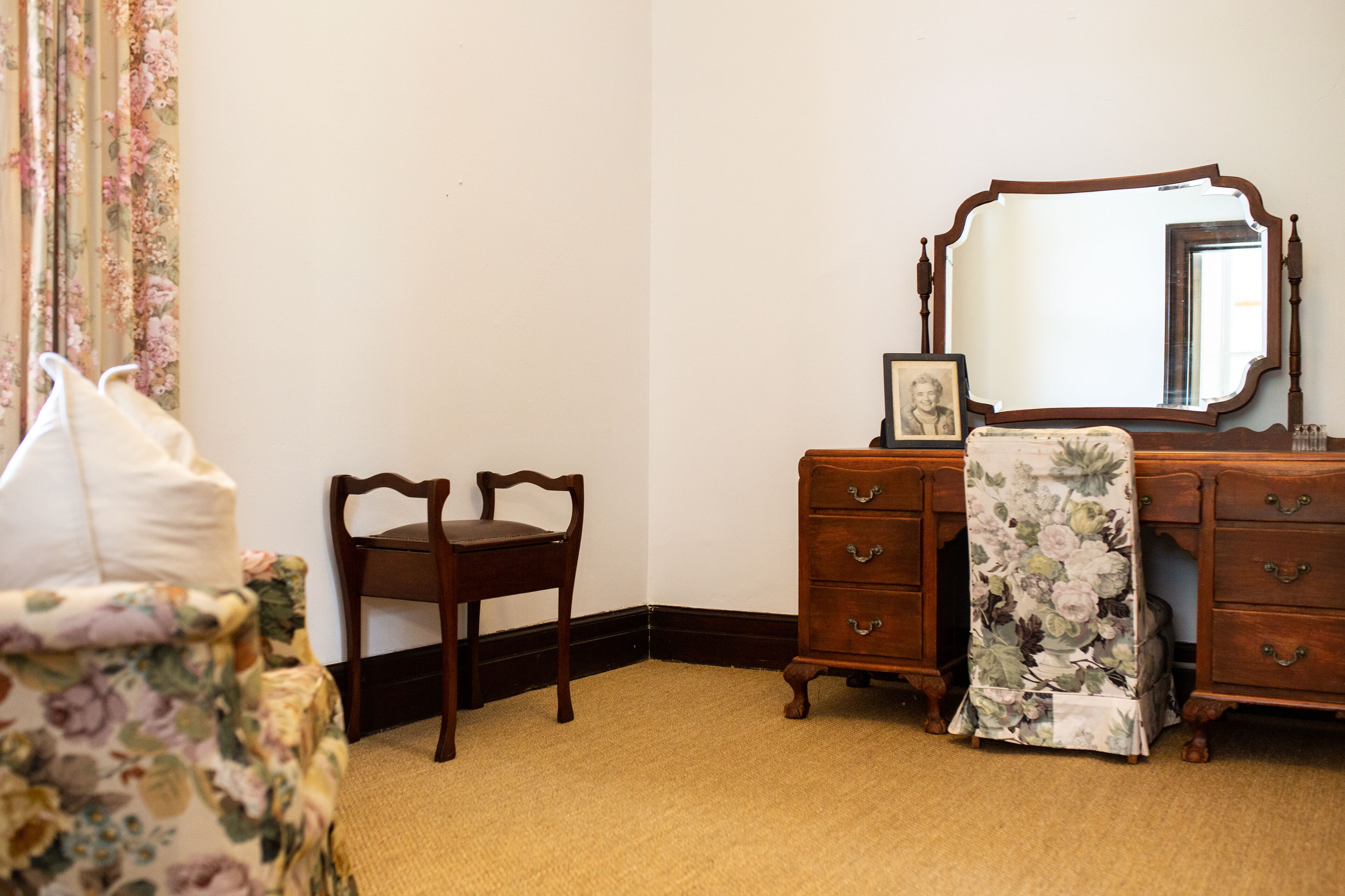 A bedroom with a wooden dresser, a chair, a bed with a floral bedspread, and a large mirror.