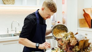 A person in a black shirt and apron preparing food in a kitchen with white shelves and a vase of flowers. - Mount William Station (Willaura)