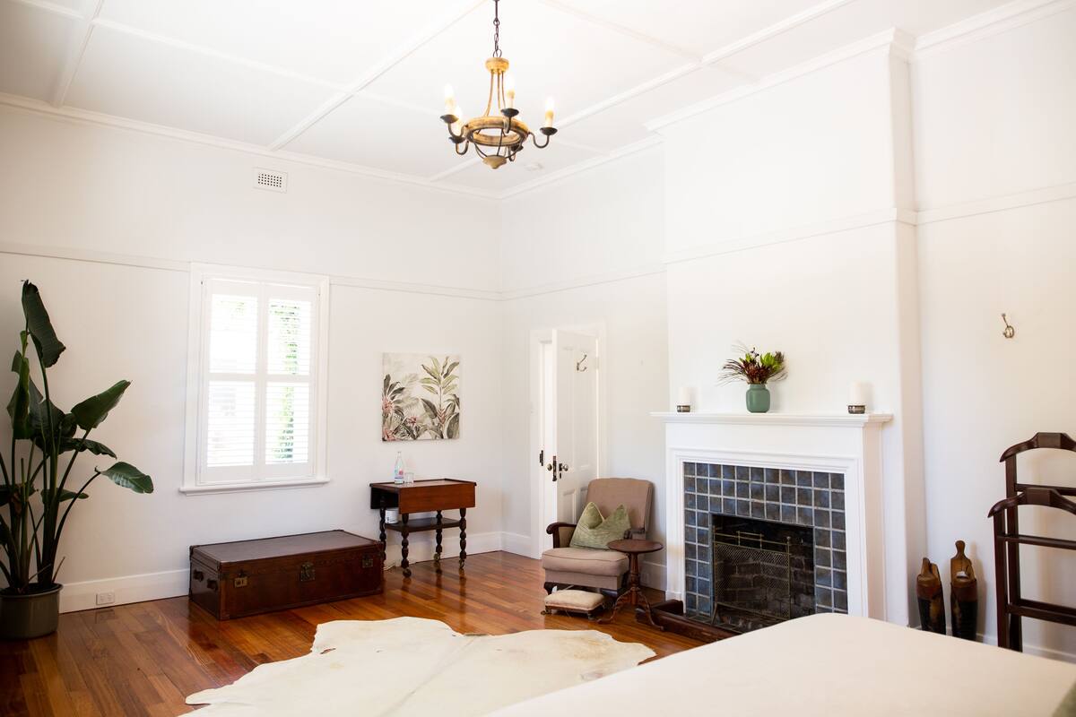 A spacious room with hardwood flooring, a large window, a fireplace, a chandelier, and a potted plant.