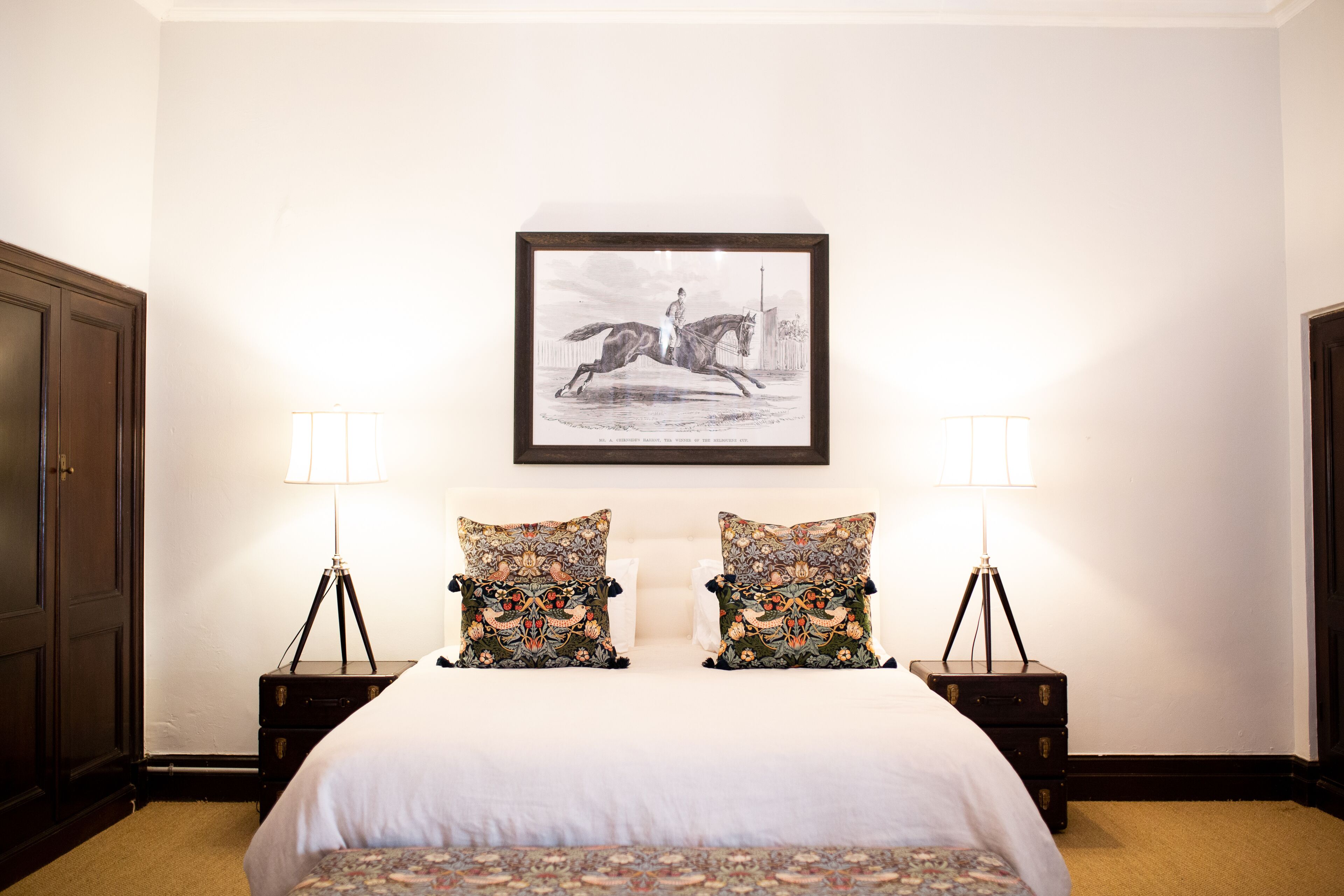 Una cama bien hecha con edredón blanco y dos almohadas decorativas, flanqueada por dos mesitas de noche con lámparas, en una habitación con un cuadro en la pared.