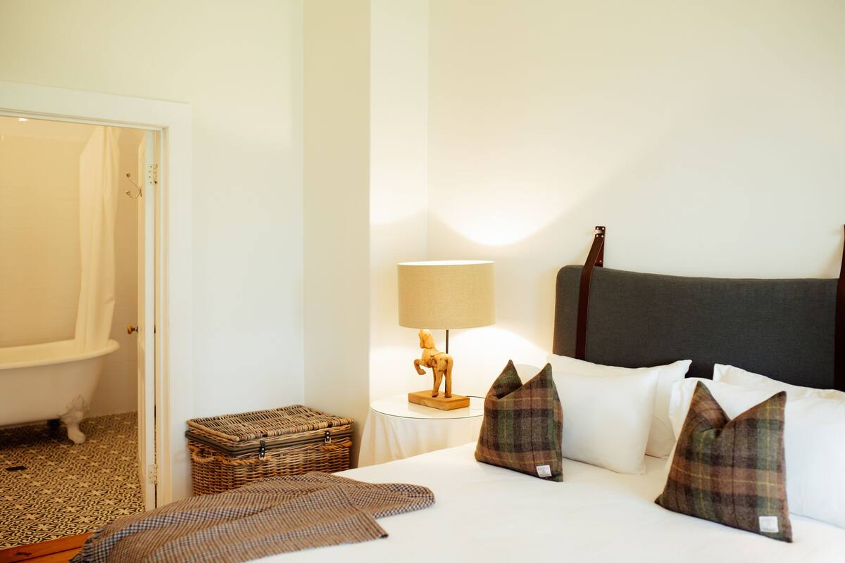 A hotel room with a bed, a nightstand with a lamp, a wicker basket, and a bathroom with a bathtub.