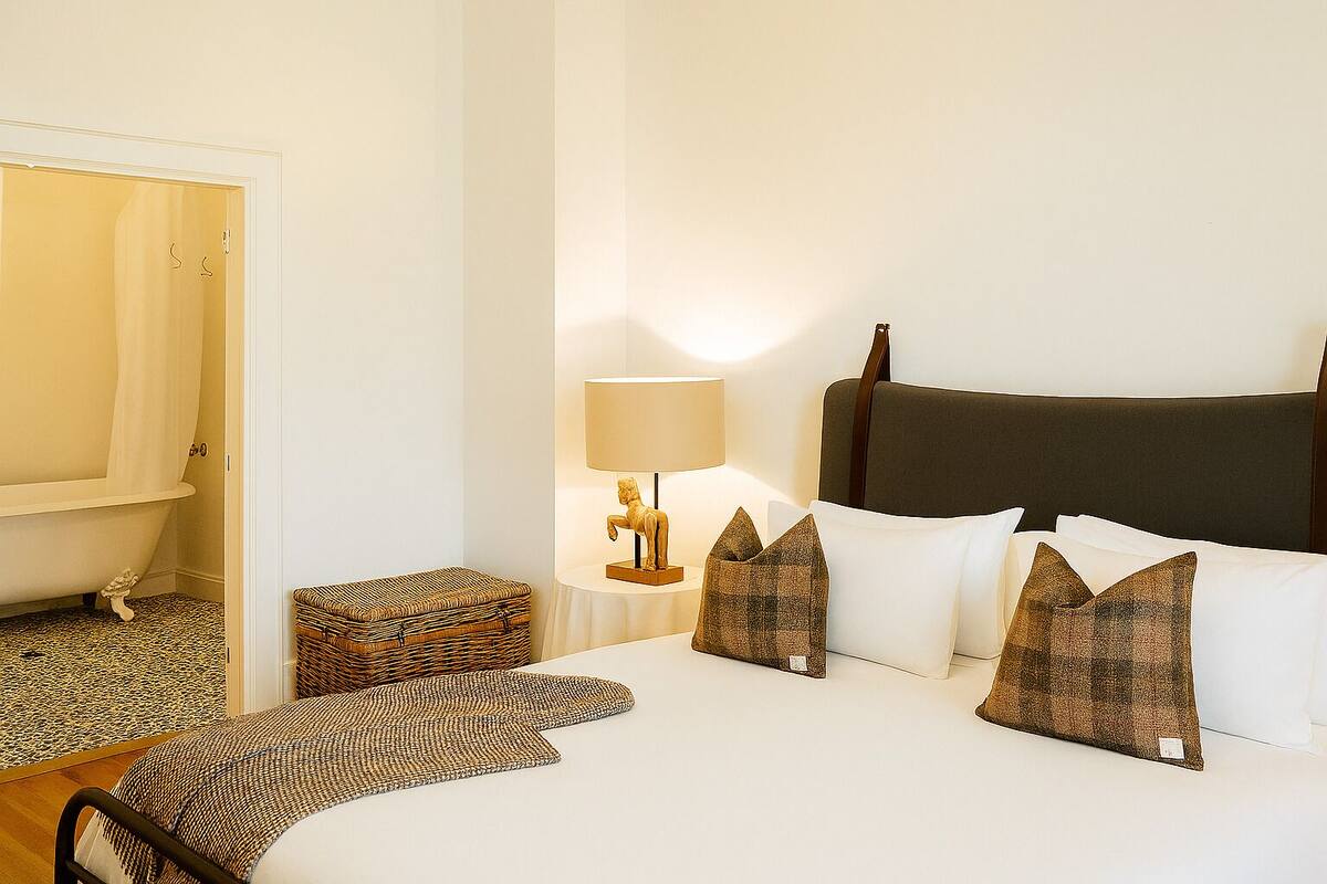 A hotel room with a bed, a bedside table with a lamp, a wicker chest, and a chair.
