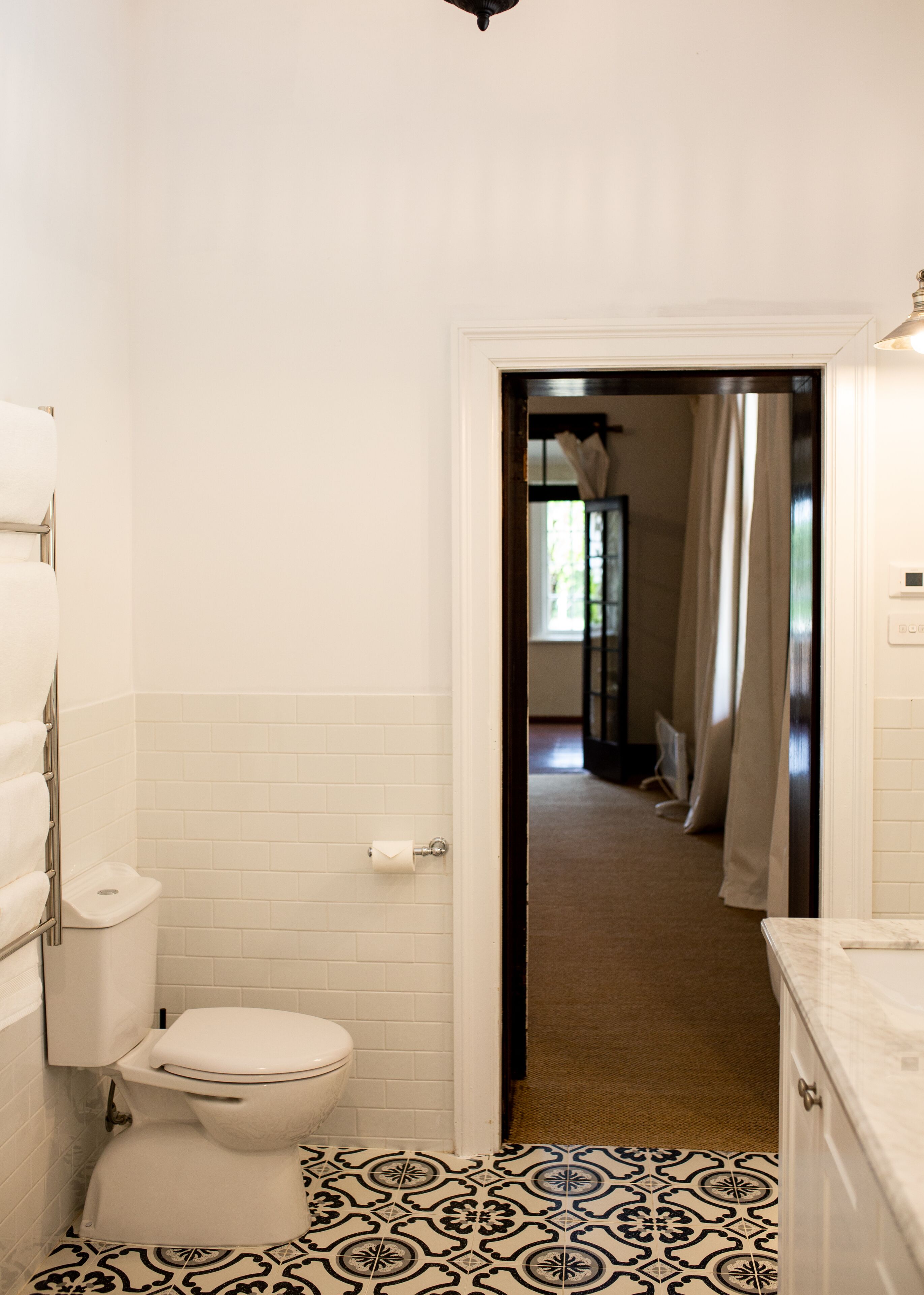 Une salle de bain avec des toilettes, des murs recouverts de carreaux blancs, un sol carrelé à motifs et une porte donnant sur une autre pièce.