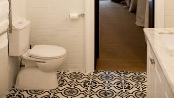 Une salle de bain avec des toilettes, des murs recouverts de carreaux blancs, un sol carrelé à motifs et une porte donnant sur une autre pièce.