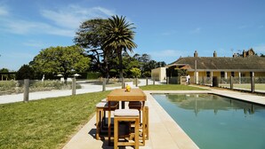 A poolside area with a wooden table and chairs, a palm tree, and a building in the background. - Mount William Station (Willaura)