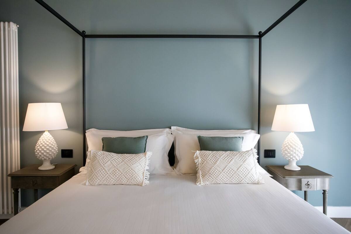 Una cama bien hecha con sábanas blancas y dos almohadones decorativos, con dos mesitas de noche y lámparas a cada lado.