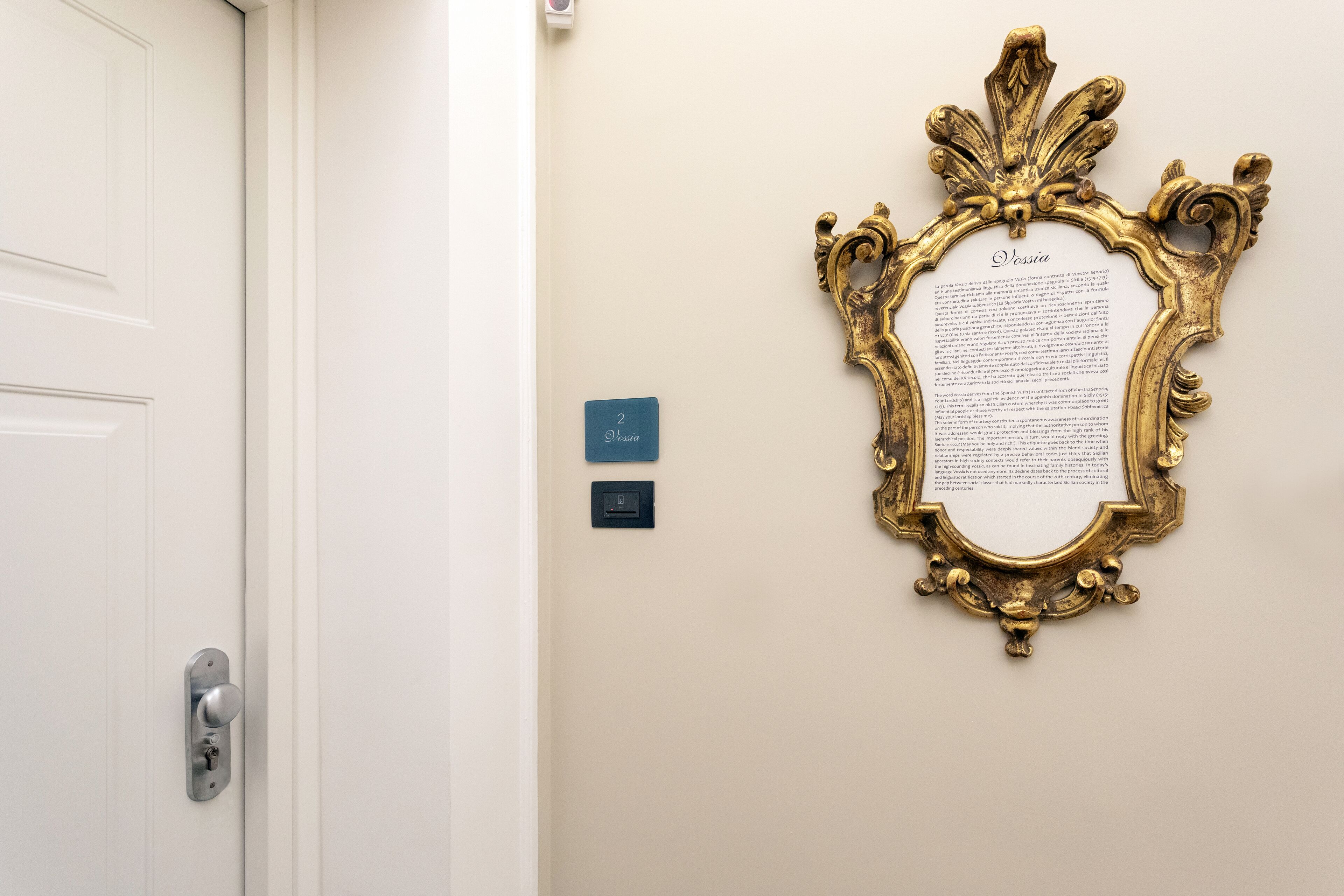 Una porta con una maniglia moderna, uno specchio con cornice dorata e una piccola targa blu appesa al muro.