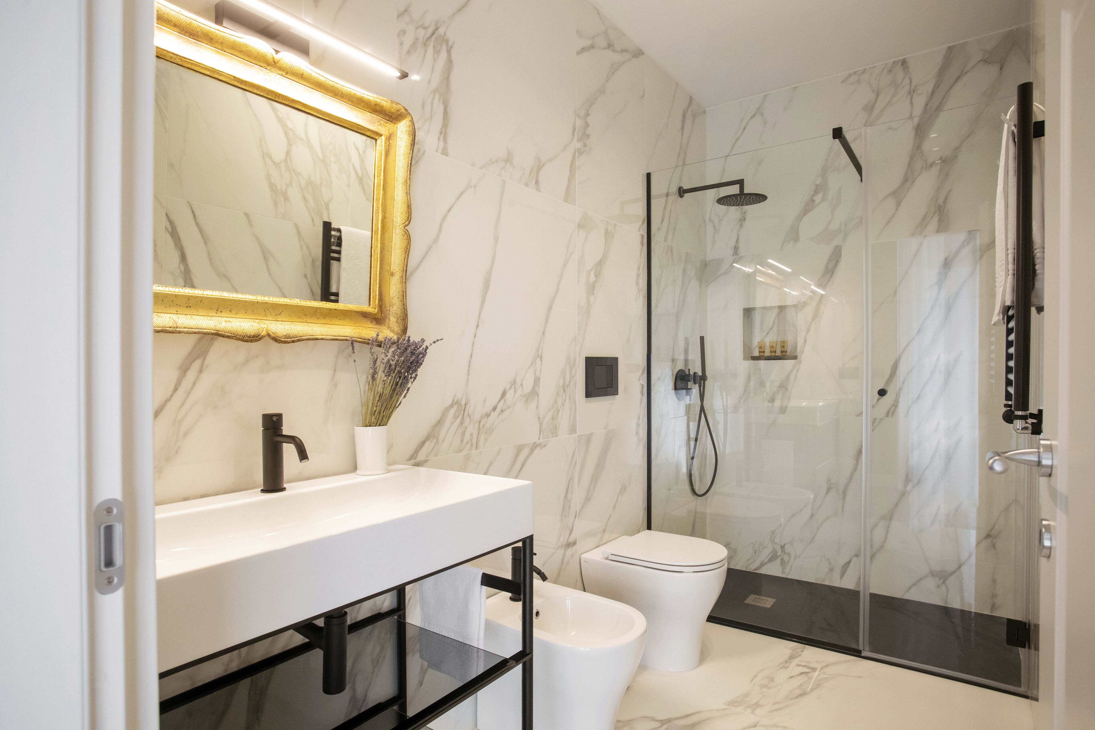 Un baño moderno con pared de mármol, un lavamanos blanco, un grifo negro, un inodoro blanco y una zona de ducha con mampara de cristal.