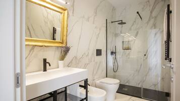 Un baño moderno con pared de mármol, un lavamanos blanco, un grifo negro, un inodoro blanco y una zona de ducha con mampara de cristal.
