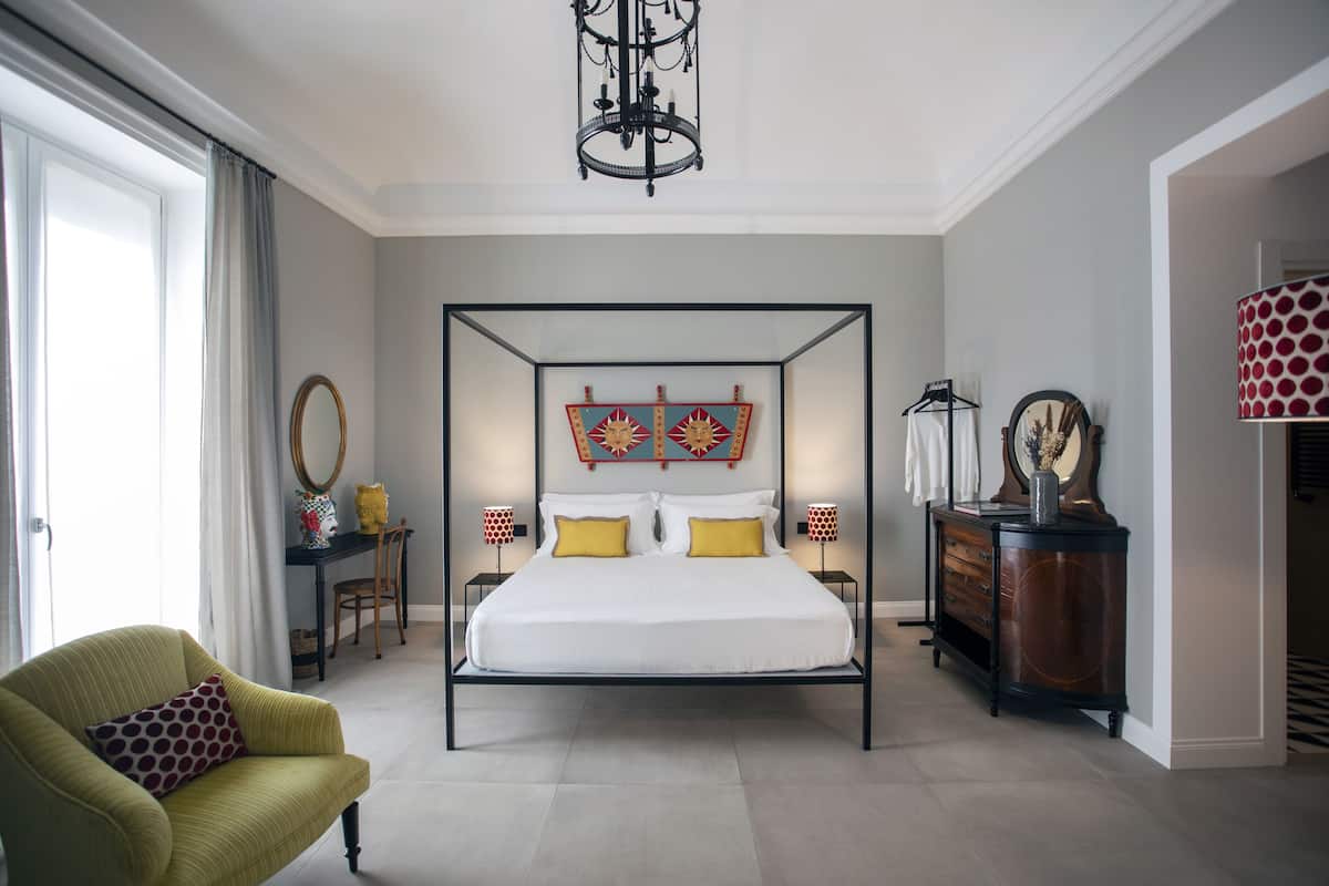 Una cama con dosel con un edredón blanco y almohadones amarillos, flanqueada por dos mesitas de noche con lámparas, un aparador de madera y un sillón verde con un almohadón estampado.