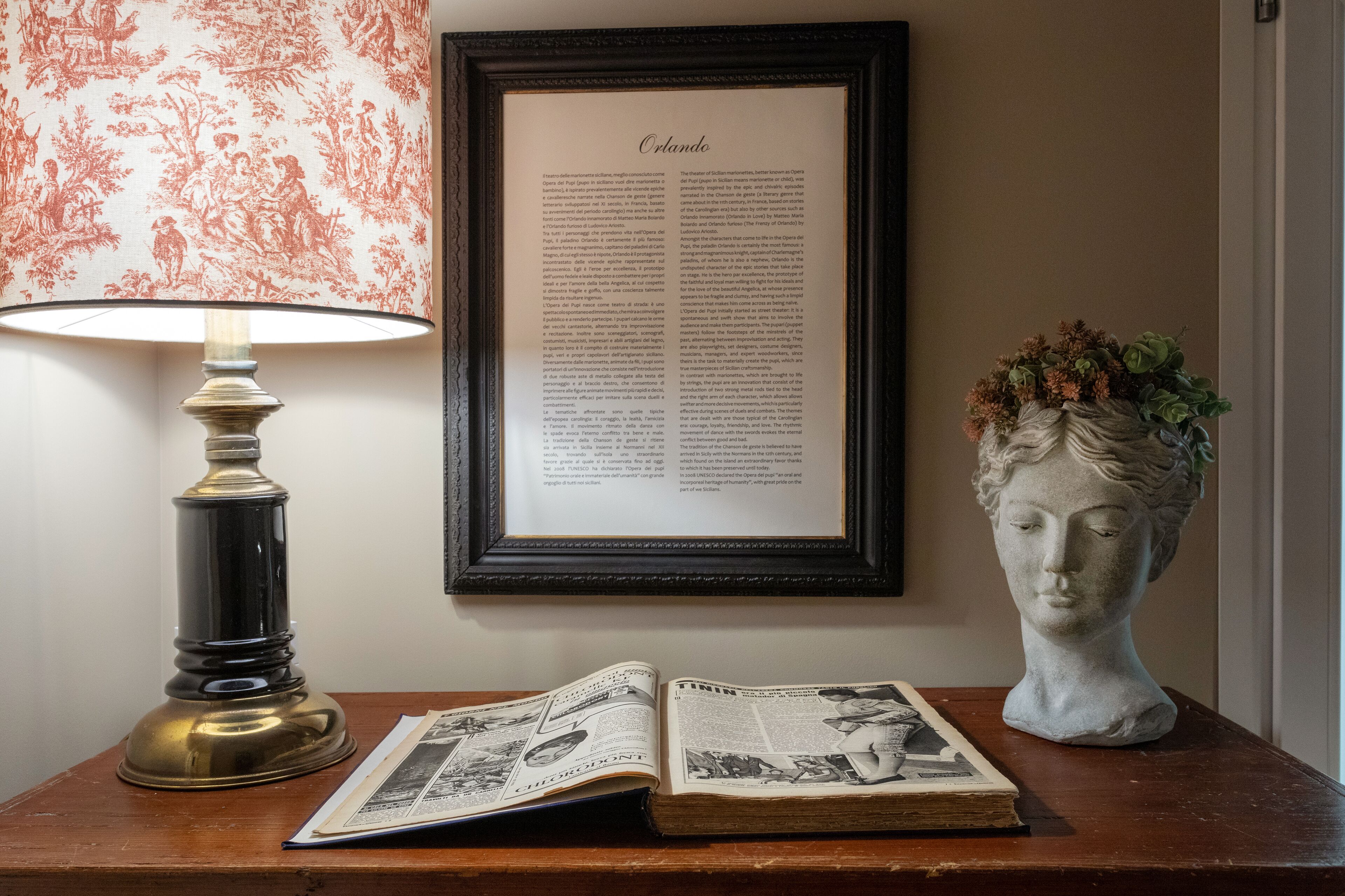 A framed text titled "Orlando" on a wall, a table lamp, an open book, and a bust sculpture.