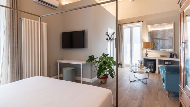 A modern hotel room with a large bed, a desk, a television, a plant, and a dining area with a sofa and lamp. - Vossia Luxury Rooms & Sicilian Living (Palermo)